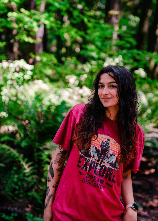 Emma (@findingarrow_) wearing a maroon Wyld Peak Explorer Tee, striking a confident pose in the lush Pacific Northwest wilderness