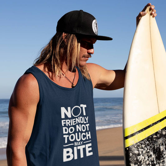 A person standing on a beach holding a surfboard wearing a navy sleeveless tank top with white text and a black cap.