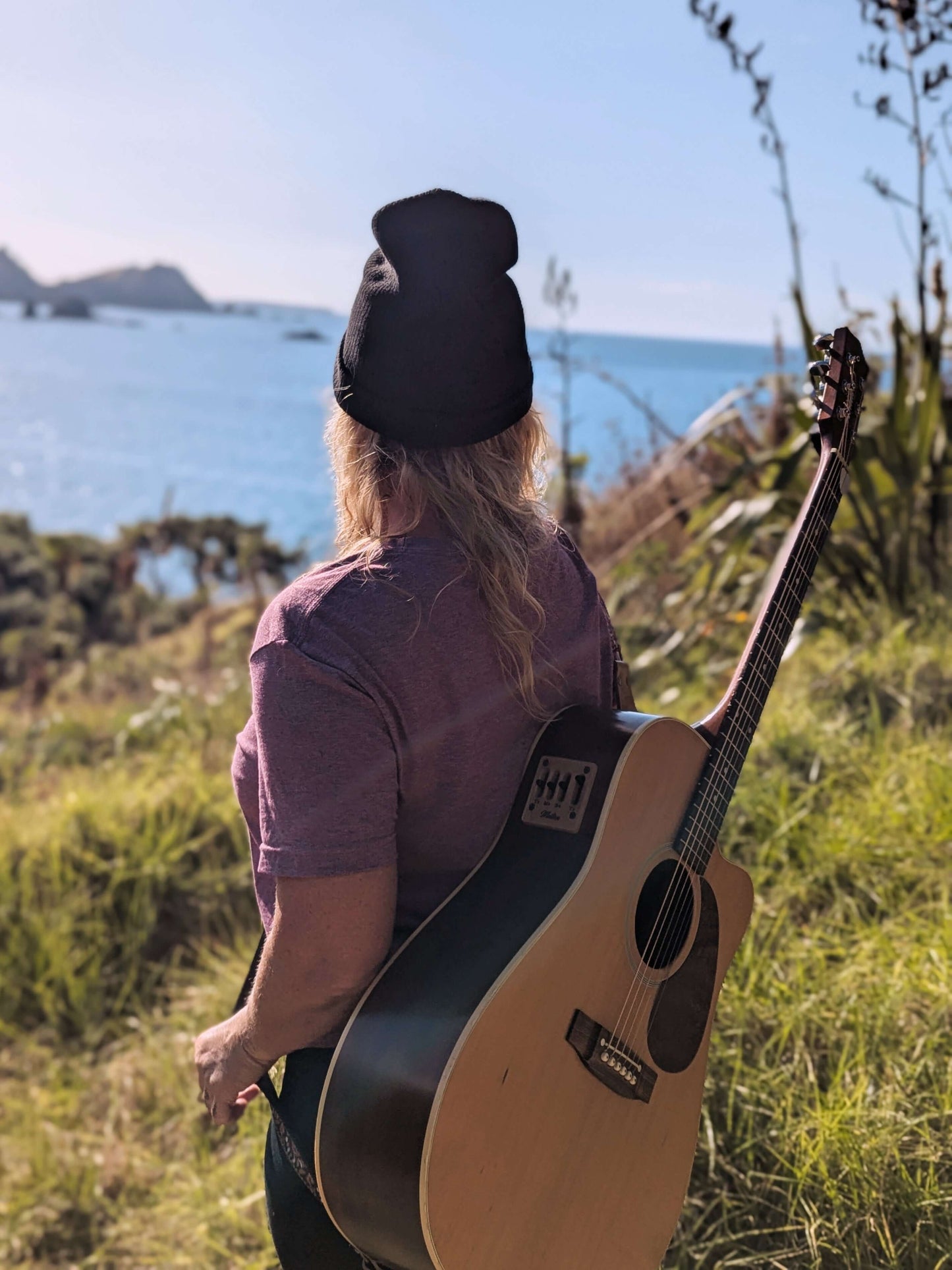 Sim Bastick at Matauri Bay in Wyld Peak maroon Explorers eco tee with guitar overlooking bay