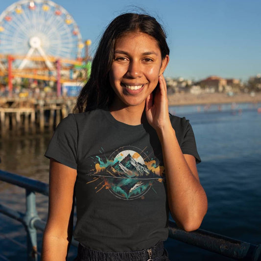 A woman smiling and wearing a black hiking t-shirt with a graphic design in the center, standing near a body of water with a ferris wheel in the background.